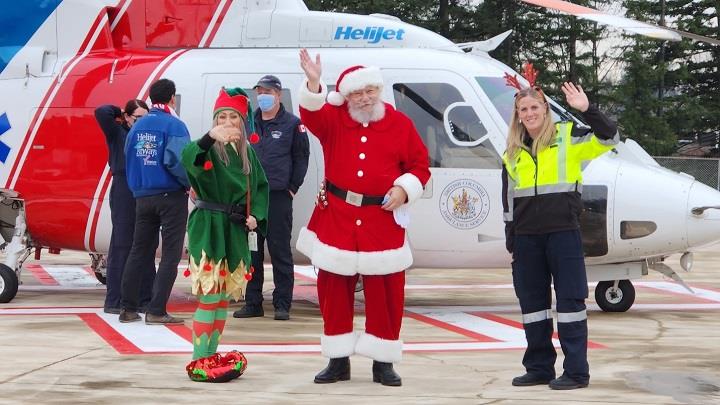An elf, Santa Claus and a paramedic waving, while standing next to a BCEHS Helijet helicopter