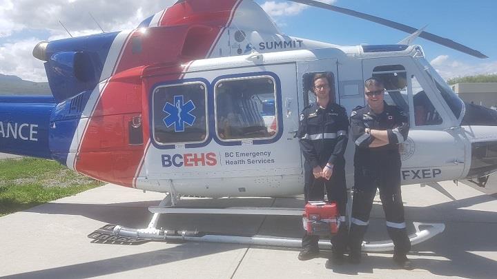 Two smiling paramedics carrying a red bag, standing by a BCEHS helicopter